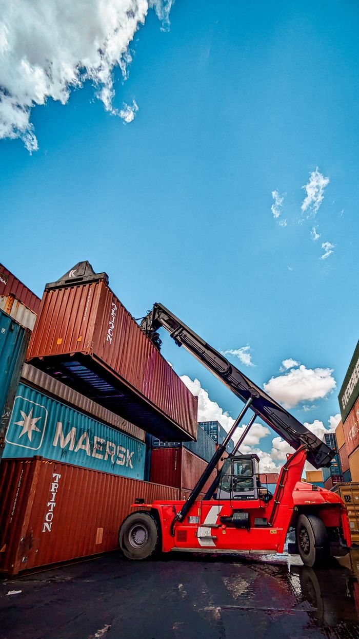 Global Markets Red hydraulic crane handling shipping containers at a port under a clear blue sky.
