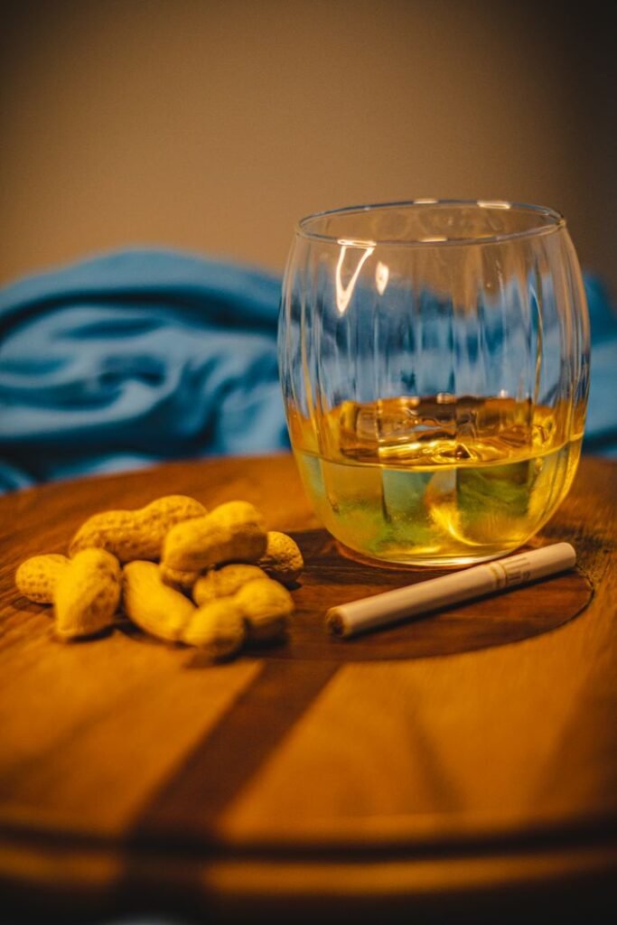 A close-up of a whiskey glass, peanuts, and cigarette on a wooden table, evoking a cozy bar atmosphere.